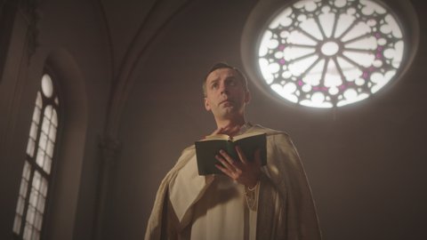 Low angle medium slowmo of Caucasian priest in long white vestment holding Holy Bible in hands while preaching sermon in dark Catholic church with round stained glass window behind him