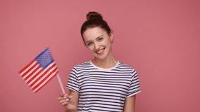 Attractive woman with bun hairstyle holding american flag, celebrating national holiday, looking at camera, wearing striped T-shirt. Indoor studio shot isolated on pink background. - Powered by Shutterstock - Get 15% off with code: PIKWIZARD15