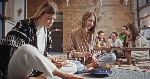 Women playing percussion instruments together. Relaxed female hippies in boho clothes playing kalimba and tank drum while sitting cross legged on floor near friends in spacious room at home