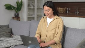 Asian young woman spending home leisure with laptop, working or studying remotel. Calm girl sitting at cozy sofa and looking at the laptop screen. Web browsing concept