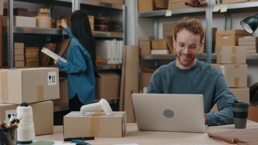 Happy caucasian entrepreneur typing parcels details at the keyboard at the laptop while his female asian colleague sorting through parcels. Small business concept | Shutterstock HD Video #1089637349