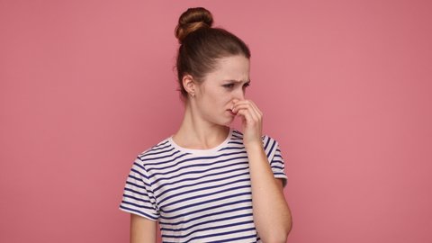 Bad smell. Young adult woman standing pinching her nose with fingers to hold breath, disgusted by stinky intolerable smell, wearing striped T-shirt. Indoor studio shot isolated on pink background.