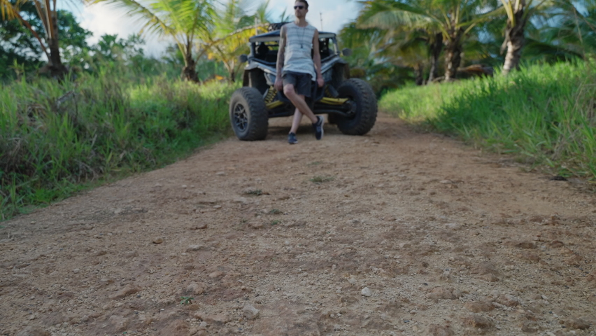 Male Wearing Sunglasses Leaning Against Bonnet Of Dirt Buggy In Empty Path In Punta Cana. Low Angle, Dolly Forward