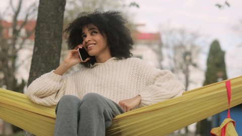 Portrait of attractive African American girl having phone talk sitting in a yellow hammock. Young beautiful curly girl in the park.
