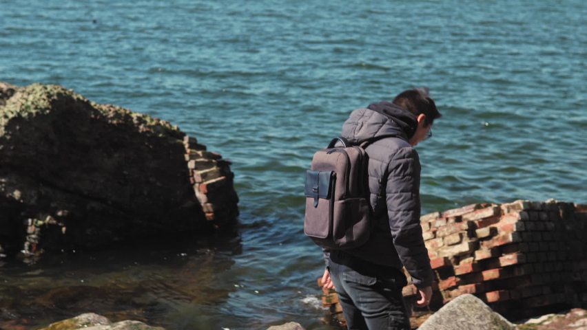 Young male tourist with a backpack over his shoulder is climbing at old abandoned ruines on the seashore and shoot a seascape into his smartphone.