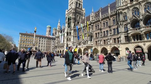 Munich, Germany 16.4.2022 People sightseeing in European city. Tourists walking in Marianplatz in front of famous Neo Gothic city hall with hanged Ukrainian flags for support. 