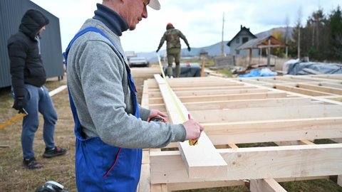 Man worker building wooden frame house on pile foundation. Carpenter using tape measure for measuring wooden planks. Carpentry concept.