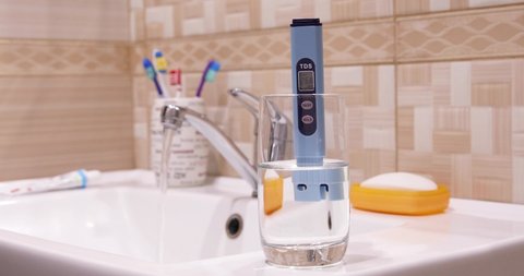 Measurement of TDS of water. Electronic pH meter in a glass of water. In the background there is a tap for drinking water, toothpaste and toothbrushes, hand soap.  Water flowing from the tap