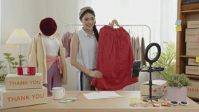 Asian young woman blogger showing clothes in front of smartphone camera while recording vlog video and live streaming at her shop. SME or freelance business online and delivery concept.