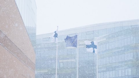 Finnish flags and European Union flag on white flagpoles against the modern building during the strong snowfall in Helsinki, Finland 