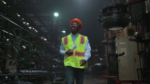 Black hair supervisor walking tyre warehouse wearing modern safety uniform. African american man industrial engineer holding paper factory plan at manufacturing company. Attractive man facility work. 
