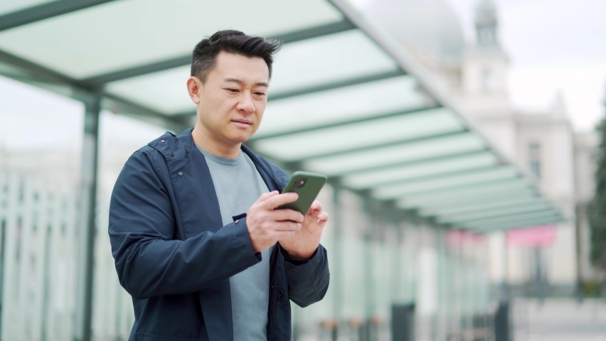 Senior Chinese tourist uses mobile phone app to book accommodation and call taxi, man near airport with suitcase. Asian man using smartphone app to rent a car at the bus stop. waiting public transport Royalty-Free Stock Footage #1089660495