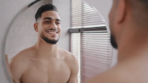Close up male reflection in mirror handsome 30s unshaven confident arabian indian bare naked man in bath smiling toothy sexy shirtless bearded millennial hipster guy happy smile getting ready to date