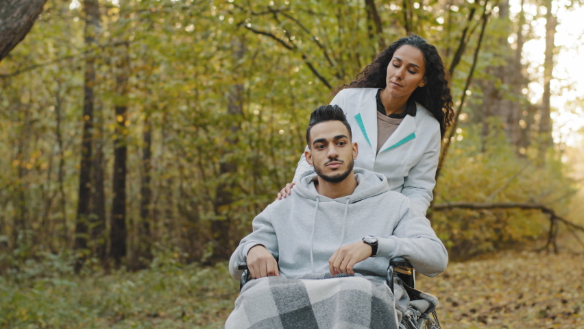 Smiling nurse girl in medical gown pushing young guy in wheelchair walking in hospital park enjoy walk in fresh air communicate feel happiness woman taking care to patient man health rehabilitation Royalty-Free Stock Footage #1089662489