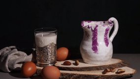 Pouring milk from a clay decanter into a glass goblet. Eggs and nuts are laid out on the table
