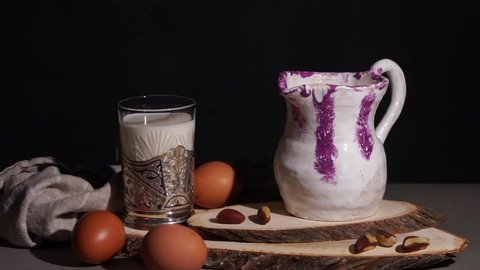 Pouring milk from a clay decanter into a glass goblet. Eggs and nuts are laid out on the table