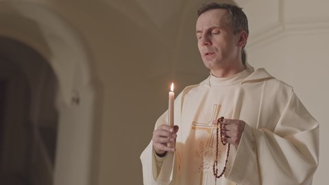 Medium slowmo of mature Caucasian priest in long white robe blowing out candle after pray, holding rosary beads in hands