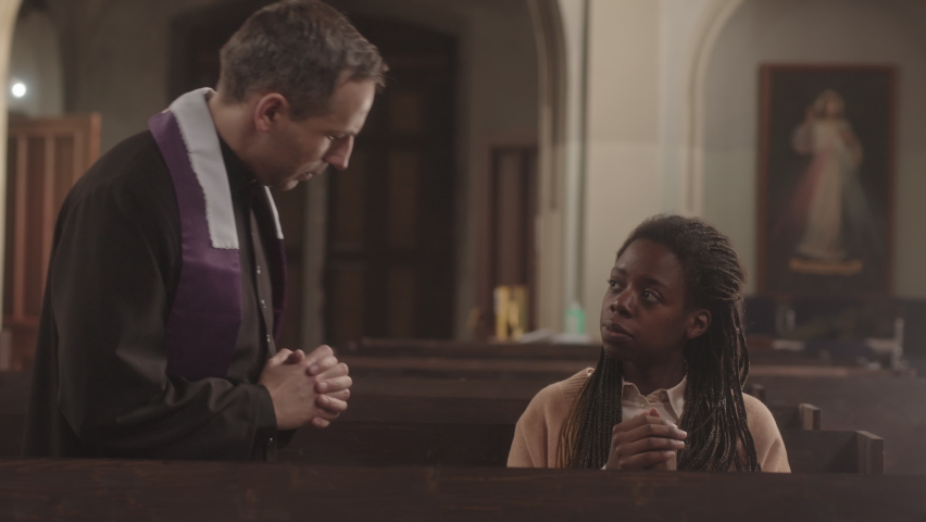 Slowmo of mature Caucasian clergyman and young African American female parishioner talking then starting praying with their hands folded in prayer, sitting together on wooden bench in Catholic church | Shutterstock HD Video #1089666649