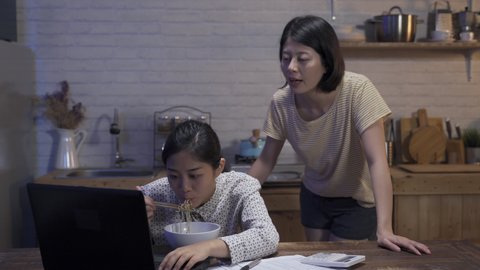chinese woman is helping check and pointing at the data on the document as her roommate is working on the computer and having supper at home.