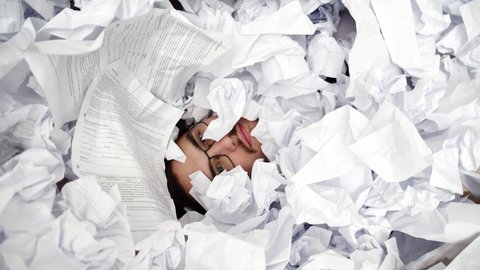 Face of male office worker screaming buried under pile of crumpled papers, top view. Man screams from under pile crumpled papers