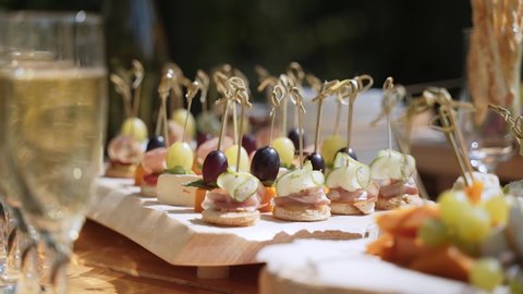 Wasps fly over canapes with skewers stuck in, neatly arranged on stand on wooden table in sunlight. Nearby are filled glasses of champagne with bubbles.
