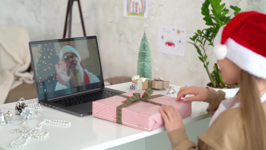A young girl with a Santa hat greeting Santa Claus during an online call for Christmas (New Year) with congratulations. Child talking with Santa. Coronavirus pandemic during the winter holidays.