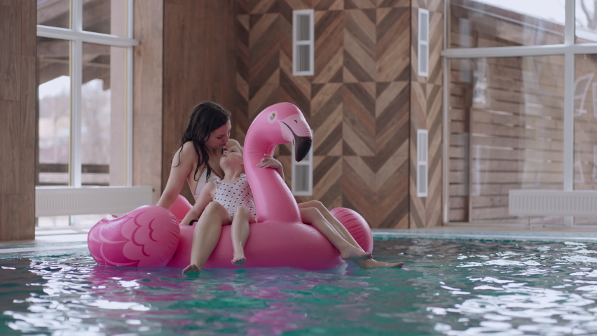 careful mother and cute little girl are floating on inflatable flamingo in swimming pool, family in water park