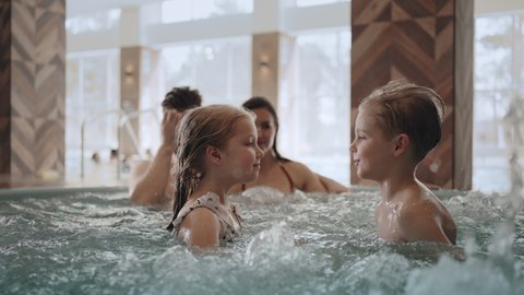 little boy and girl are playing in pool, family is resting in jacuzzi of modern spa center