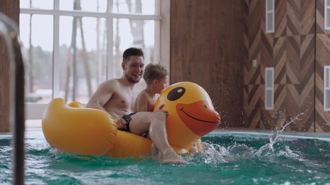 cute little boy and his dad are riding funny inflatable duck in swimming pool, having fun and resting together