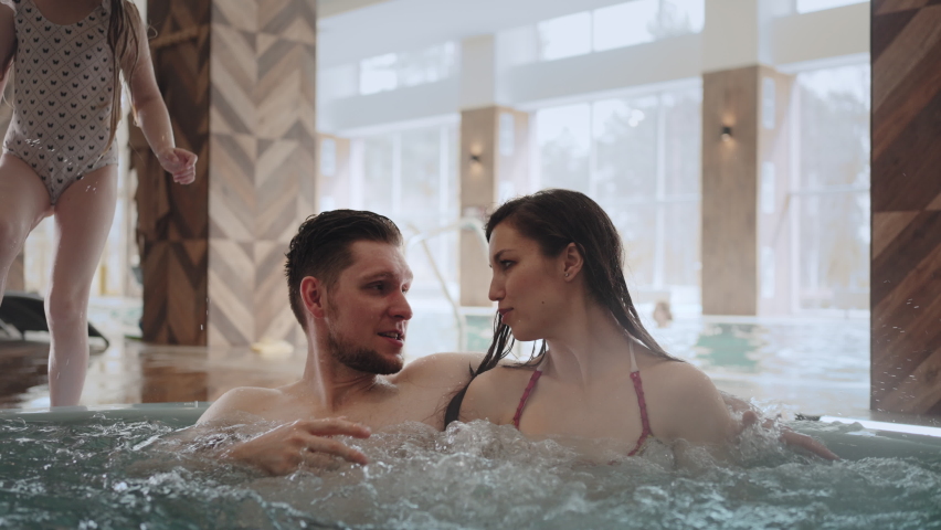 family is resting in swimming pool, parents are chilling in jacuzzi, son and daughter are playing in water