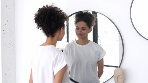 Lovely happy African American curly-haired girl in a white basic t-shirt, stands in front of a mirror in the morning, admires herself, smiles, starts the day well, good mood, looks at her face