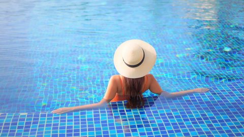 Woman in orange swimsuit and straw cap relaxing in clear warm water of luxury resort swimming pool. Lady sunbathing and enjoying vacation. Asian girl standing in pool looking around.