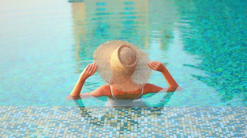 Woman wearing hat standing in warm clear swimming pool with colorful blue tile. Asian lady relaxing in resort pool stretches arms over head and takes in sunshine of tropical island paradise.