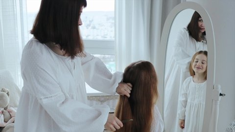 Young adult mother is combing long hair of her little daughter in front of the mirror. Mom and daughter dressed in white retro-styled nightdresses spending time together in the morning, 4k