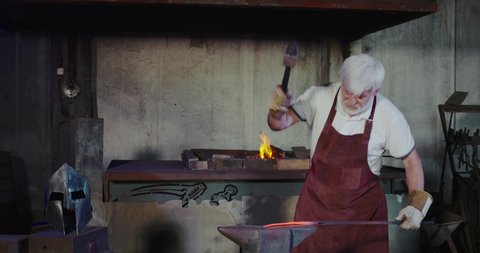 front view of old man in apron and gloves forging in smithy. Strong blacksmith with grey hair and beard long stick from furnplace taking, with hammer beating. Concept of forging.