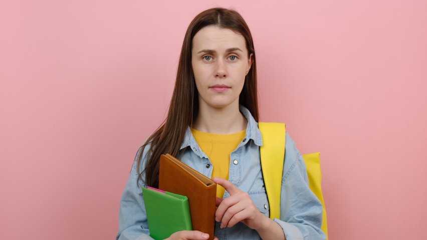 Portrait of confused fun girl teen student hold books spreading hands say oops ouch oh omg i am so sorry, wears shirt and yellow backpack, posing isolated over pink color background wall in studio Royalty-Free Stock Footage #1089691961