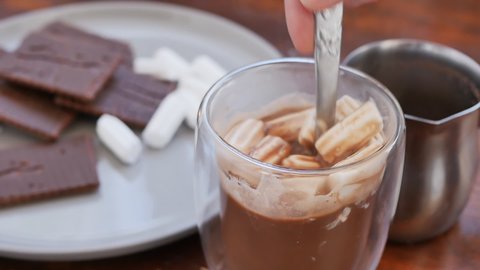 a woman's hand stirred cocoa and marshmallows in a glass glass with a spoon and took the glass from the table. warm cocoa milk drink with chocolate chip cookies in the evening. What could be better