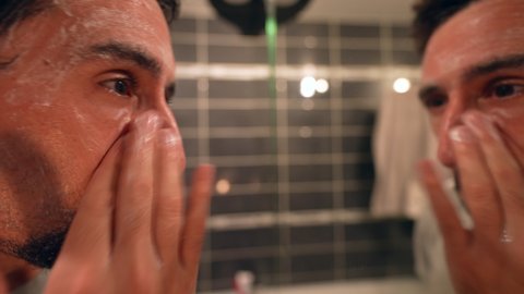 Man applying lotion cleanser washing face in front of mirror