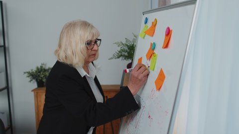 Senior business woman manager making notes on white board with stickers analyzing infographics using laptop computer sits at workplace desk. Mature old grandmother freelancer working at home office