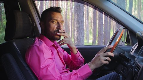 Businessman smoking a cigarette while driving, looking at the camera and talking. A young man in a car on a background of green trees, a hot sunny summer day. Warm soft light, close-up. 4K UHD.