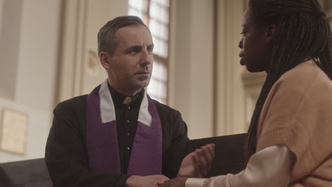 Waist up slowmo of mature Caucasian priest in black and purple robe and young African American female parishioner having conversation, sitting on wooden pew in Catholic church holding hands