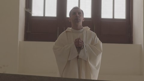 Medium slowmo of mature Caucasian priest in long white alb praying to God and crossing standing by altar with rosary beads in hands