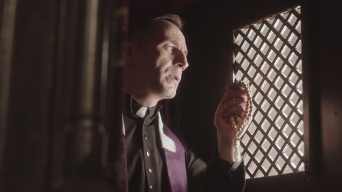 Waist up slowmo of mature priest wearing black and purple robe talking to parishioner while sitting in dark confession booth with rosary beads in hands