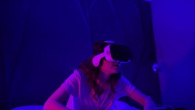 Young Woman Uses Virtual or Augmented Reality Glasses Sitting On A Bed in Dark Room, Female VR Headset User. Entertainment leisure time