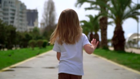 Child girl runs away to park. Kids walk outside on street. Happy childhood concept. A little Caucasian girl walks forward with her back. Walk on a sunny day. Summer walk among green trees and palms.