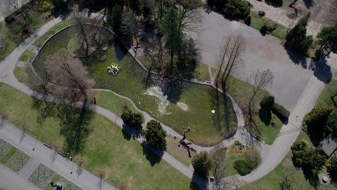 Park in Olomouc with a pond reflecting the spring sun on the surface. Fountain and bathing ducks. The green flora of trees, groves and shrubs led by sidewalks. Czech Republic. Drone view
