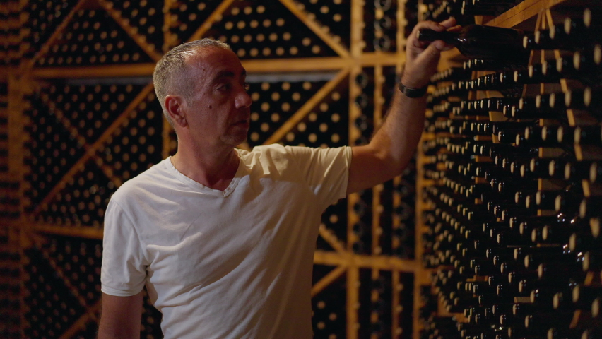 Focused grey-haired man choosing bottle of high-quality wine in slow motion. Medium shot portrait of Caucasian winemaker standing in wine cellar selecting alcohol beverage. Luxury and manufacturing Royalty-Free Stock Footage #1089709743
