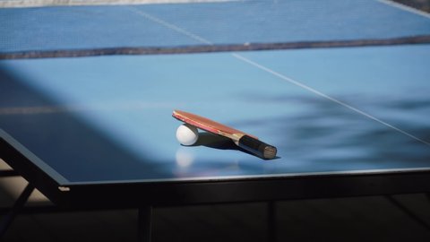 Old shabby table tennis racket with rewound handle lies on blue tennis table, covering white ball. Sunlight and shadow from tree branches hit foldable training table with net.