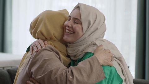 Smiling happy young woman in white hijab visiting her mother. Adult daughter in hijab kissing her mother, celebrating Eid or Mother's Day. Young woman and her mother hugging each other with happiness.