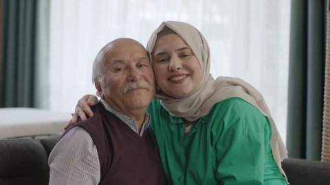 Smiling happy young woman in hijab visiting her elderly father. Adult daughter in hijab kissing her father, celebrating Eid or Father's Day. They are smiling and looking at the camera.